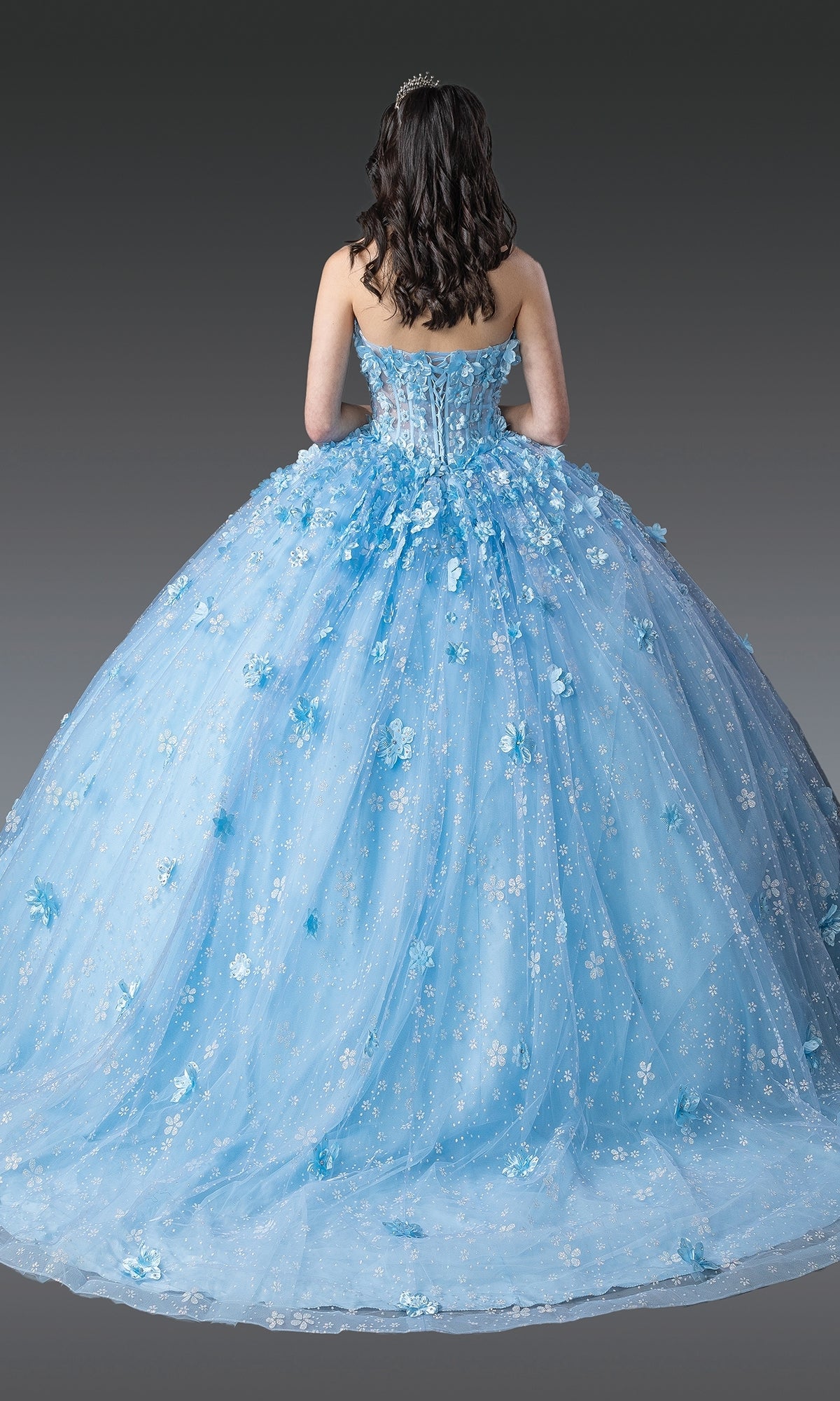 A young woman in the Blue Quinceanera Dress 1972 By Dancing Queen, featuring a strapless light blue design with 3D flowers and a tiara, stands facing away against a gray background.